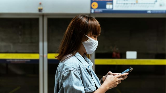 Asian woman wearing a face mask, texting on smartphone, indoors at railway station.