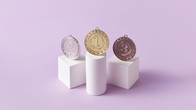 Gold, silver, and bronze medals displayed on white pedestals with a pink background.