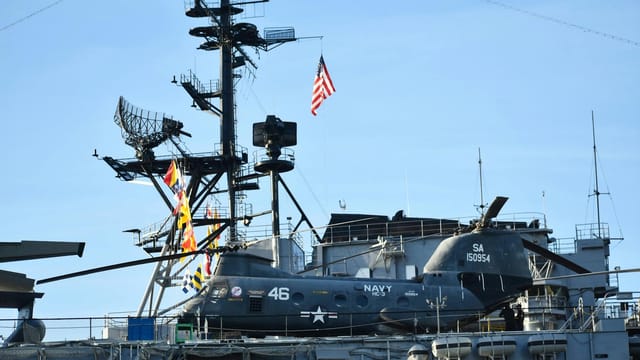 A US Navy helicopter is stationed on the deck of an aircraft carrier with radar equipment in the background.