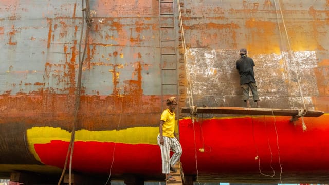 Two workers refurbishing a rusty ship hull with bright paint, showcasing industrial shipyard labor.