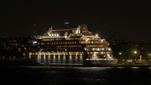 Illuminated cruise ship docked at night with city lights in the background, reflecting on water.