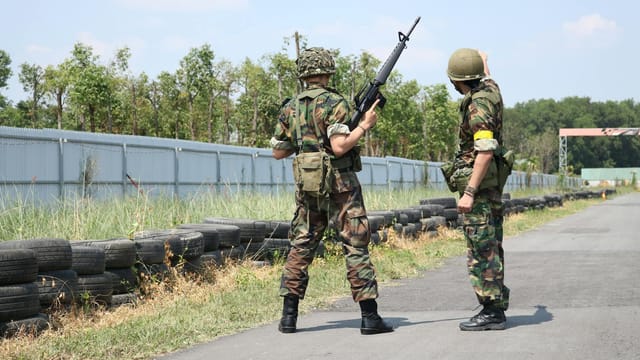 Two soldiers in camouflage uniforms with weapons stand outside on a training field.