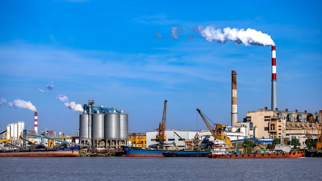 An industrial port scene with smokestacks, silos, and cranes by the water, showcasing urban industry.