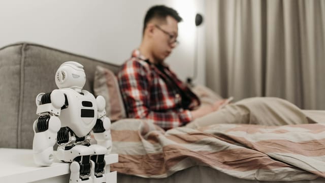 A modern bedroom featuring a robot on a bedside table and a man sitting on a bed.