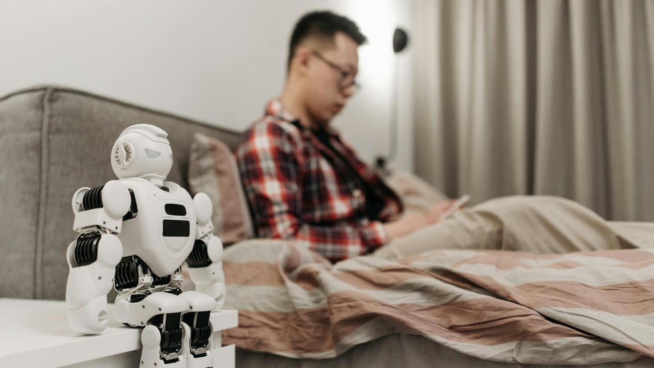 A modern bedroom featuring a robot on a bedside table and a man sitting on a bed.