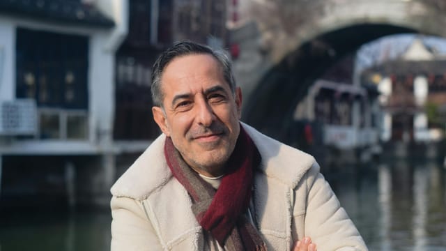 A middle-aged man smiles confidently near a canal bridge, wearing a winter coat and scarf.