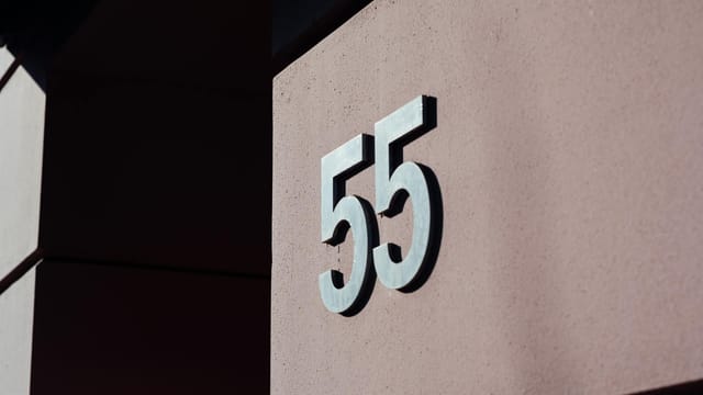 Close-up of a modern building wall with metal number 55 casting a shadow.