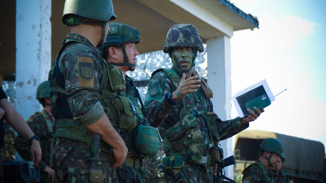 Group of soldiers in camouflage attending a briefing outdoors with tactical gear and helmets.