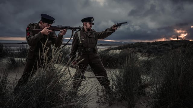 Soldiers in uniform train with weapons on a windy beach at dusk, under dramatic skies.