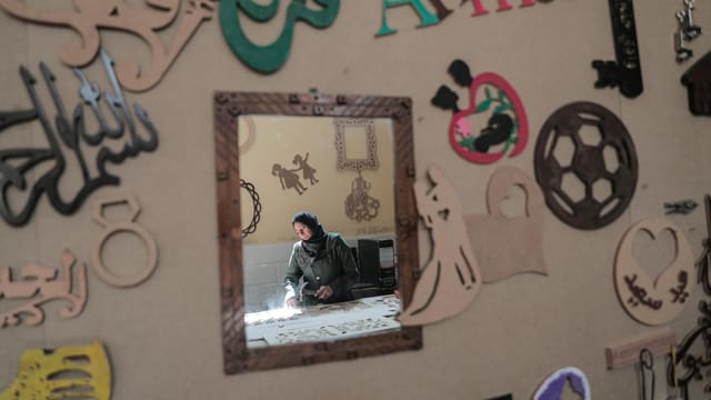 A woman in hijab reflected in a mirror surrounded by cultural decorations in a Gaza workshop.