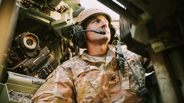 A soldier in camouflage uniform inside an armored vehicle, looking focused.