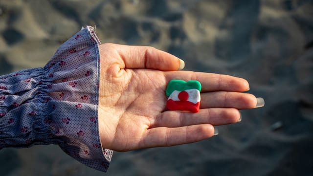 Close-up of a hand holding a colorful clay sculpture on a sandy beach with long sleeves visible.