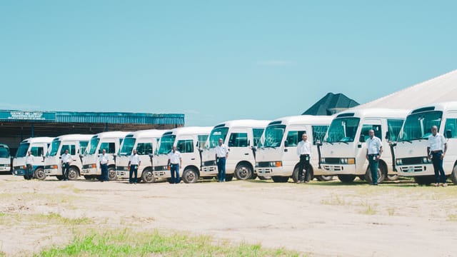 A line of minibuses with drivers standing in front, under clear skies in AK, Nigeria.