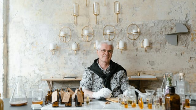 Senior male perfumer sitting among fragrance bottles in a rustic setting, creating unique scents.