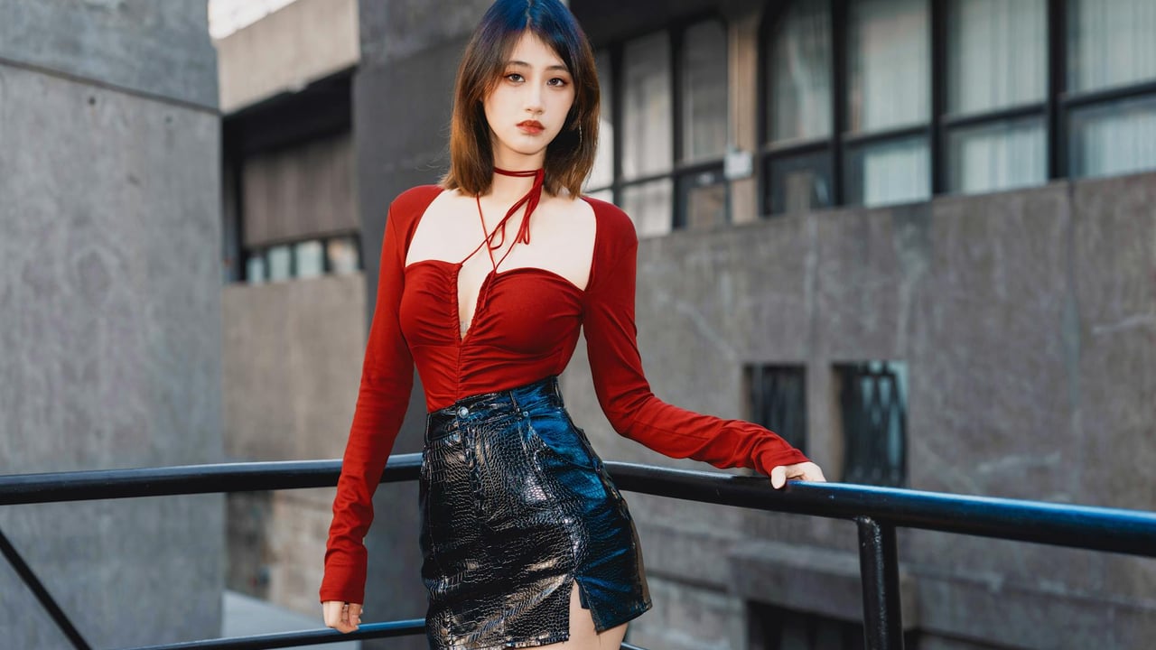 Stylish woman in red top and black leather skirt posing outdoors with modern architecture.