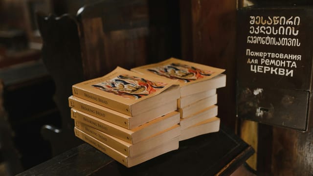 A stack of books titled 'Breaking Bread' on a wooden church surface with donation box.
