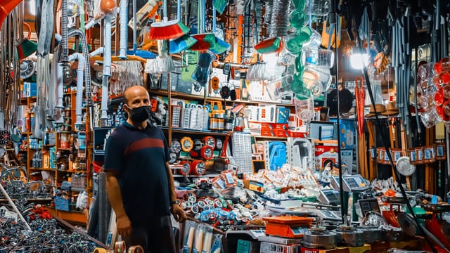 A man wearing a black facemask standing in a crowded hardware store filled with various tools and merchandise.