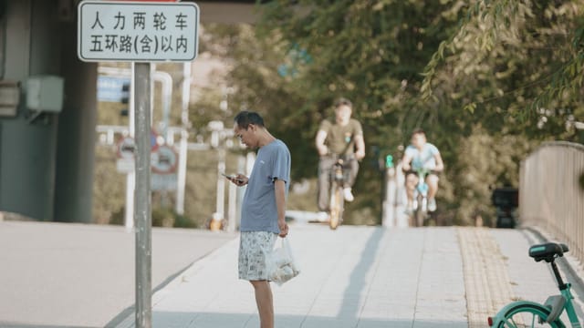 Casual man stands on a sunny sidewalk, engrossed on his smartphone, as cyclists pass by in an urban environment.