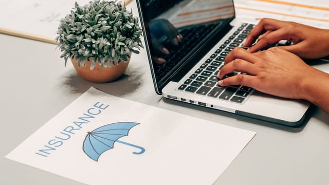 Illustrative image: Hands typing on a laptop at a desk with an insurance paper and plant, suggesting a work environment.