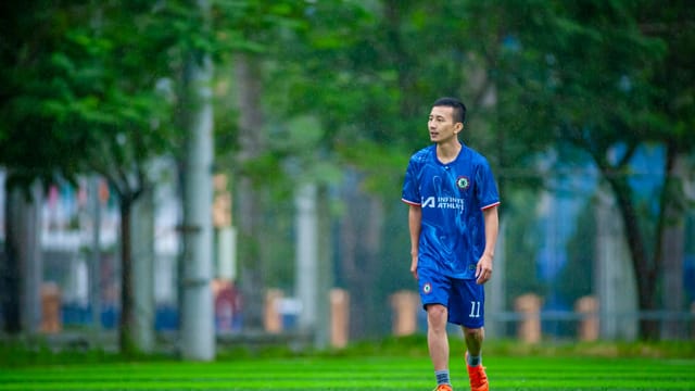 A soccer player in a blue jersey stands on a lush green field in Hanoi.