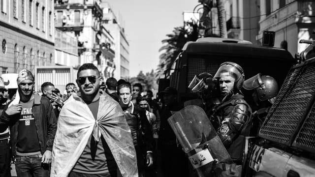 Black and white scene of protesters and police in an Algerian street, highlighting tension.