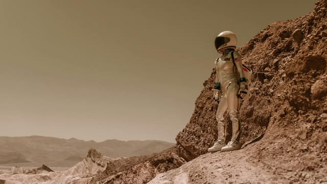 A lone astronaut in a spacesuit stands on a rugged, rocky terrain resembling an alien planet.