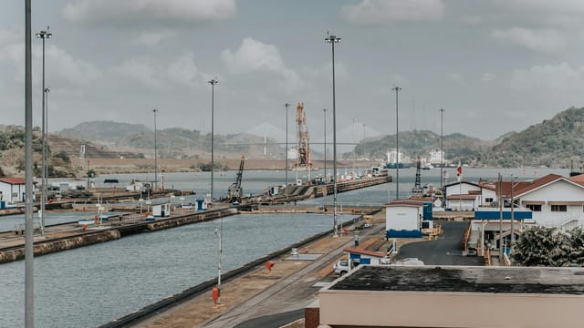 A panoramic daytime view of the Panama Canal locks with surrounding landscape and maritime infrastructure.