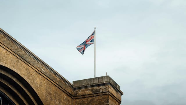 Union Jack flag waving atop a historic brick building in London, UK, against a cloudy sky.