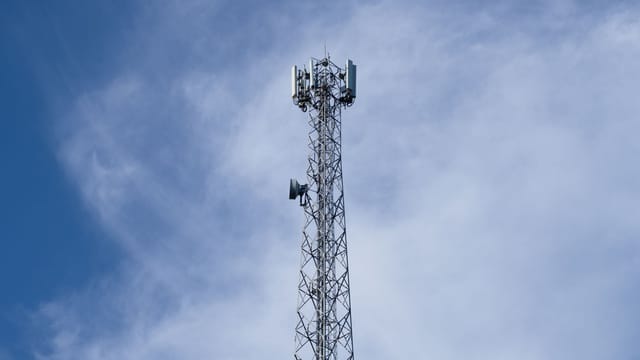 A tall metal communications tower stands against a backdrop of a clear blue sky with scattered clouds.