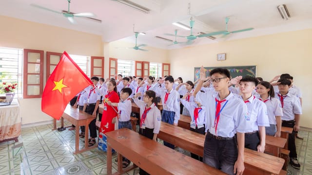 Group of Vietnamese students in uniform saluting the national flag during a classroom ceremony.
