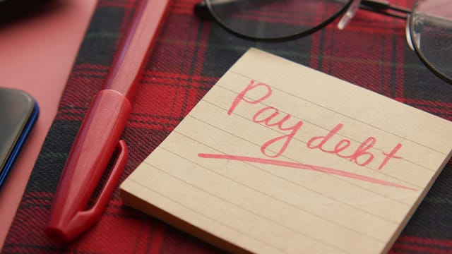 Close-up of a note reading 'Pay debt' next to a red pen on a plaid fabric, emphasizing financial reminders.