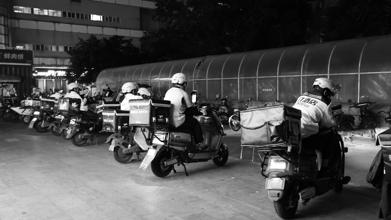 Food delivery drivers on scooters gather at night for dispatch in an urban setting.