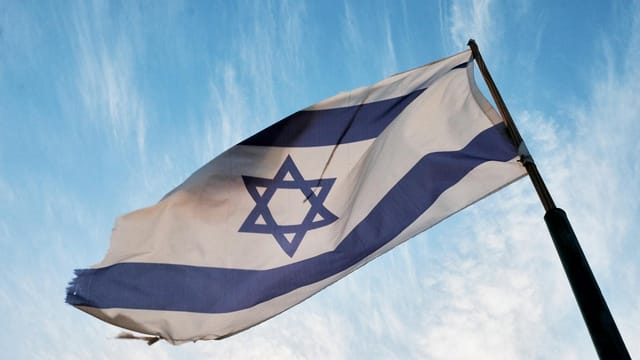 A close-up of the Israeli flag waving on a flagpole against a clear blue sky.