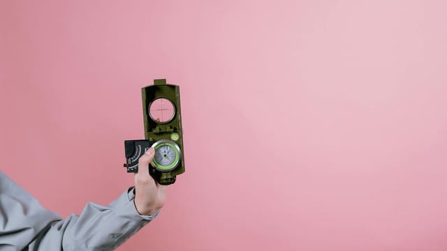 A person holds a military compass against a pink background, symbolizing navigation and direction.