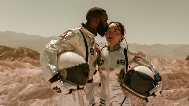 Interracial couple in space suits embracing in a desert landscape, symbolizing exploration and love.