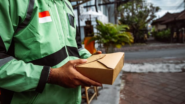 A person in a green jacket delivering a package outdoors. Urban setting, daytime.