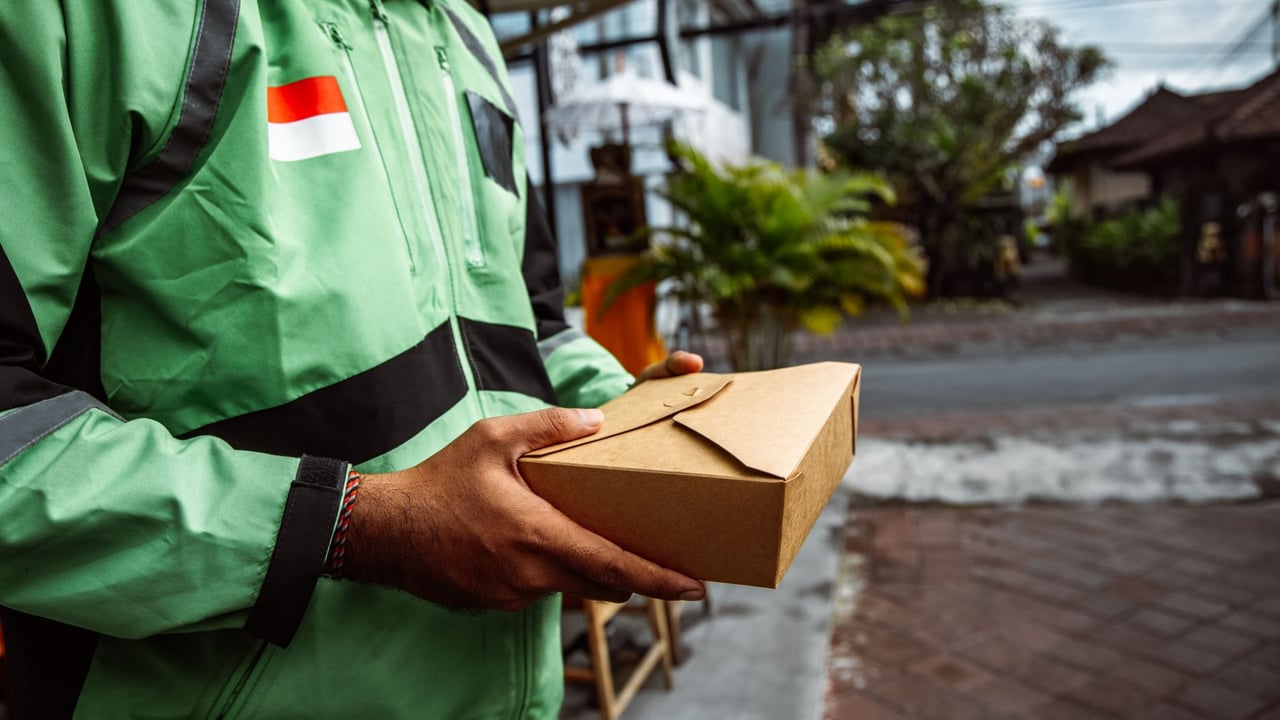A person in a green jacket delivering a package outdoors. Urban setting, daytime.