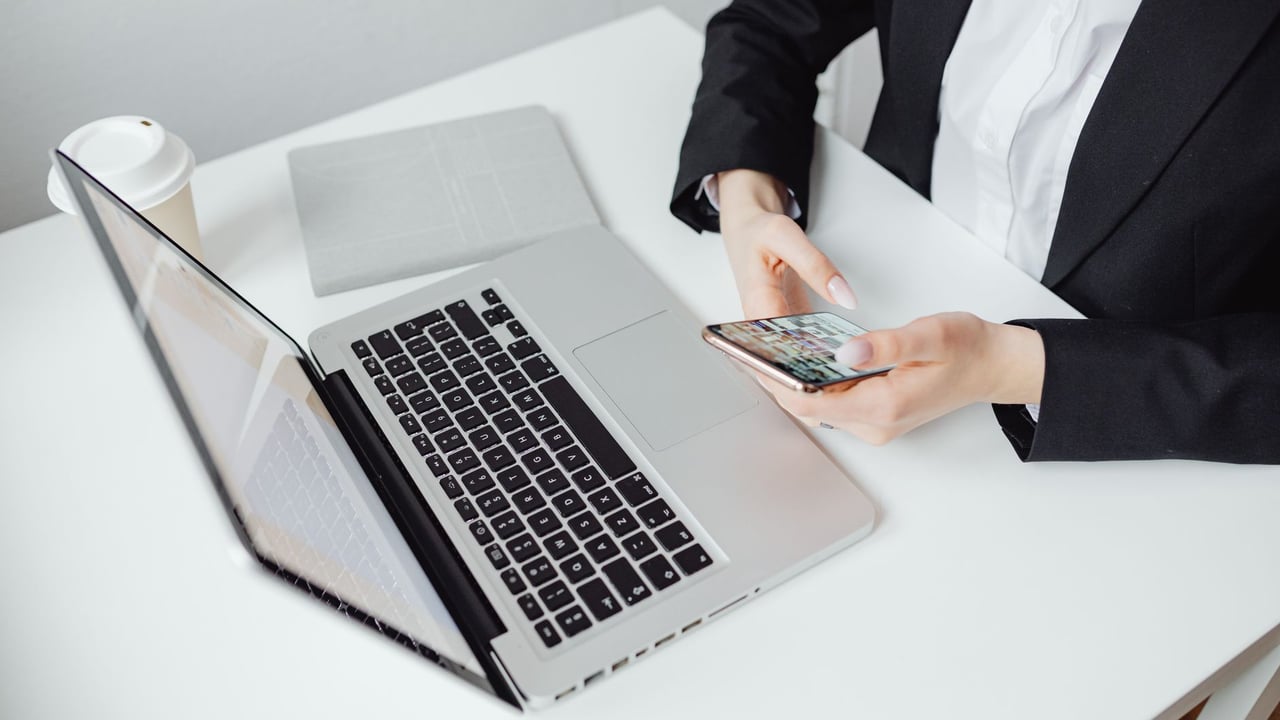 Businesswoman using smartphone at desk with laptop and coffee cup.
