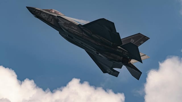 Impressive aerial view of F-35 Lightning II jet soaring through a clear blue sky with clouds.