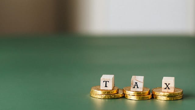 Stacked gold coins with wooden blocks spelling 'TAX' on a green backdrop.