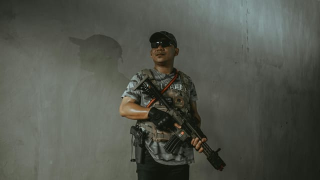 A soldier wearing tactical gear and sunglasses holds an assault rifle indoors.
