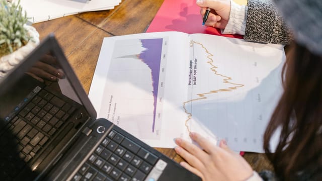 Person reviewing financial graphs and charts while taking notes beside a laptop.