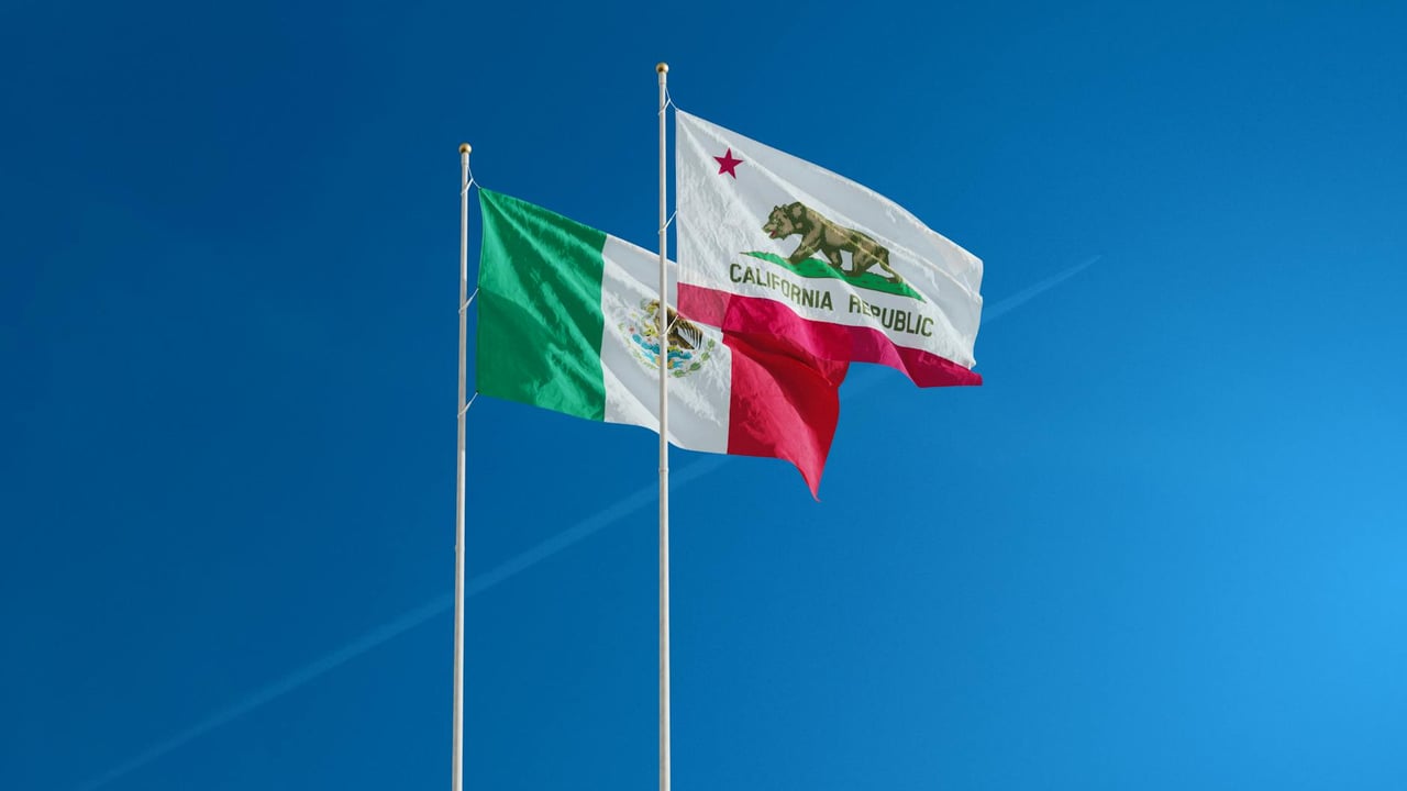 Mexican and Californian flags waving on poles against a clear blue sky.