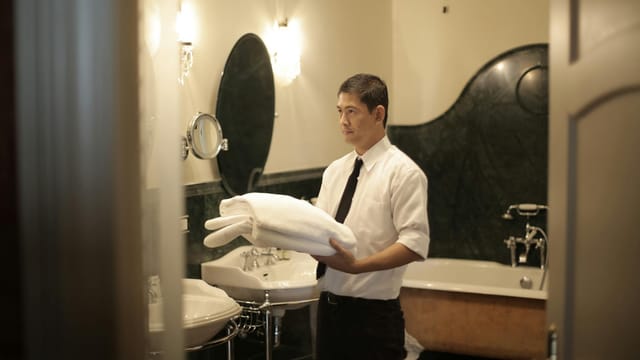 Side view of young male employee holding stack of white towels while standing in bathroom of luxury hotel