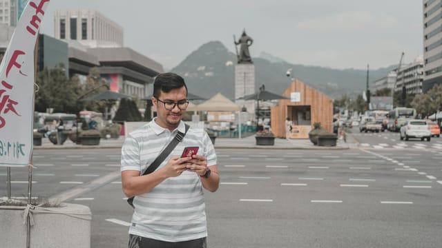 A man in a striped shirt standing on a street using a smartphone with a city skyline in the background.