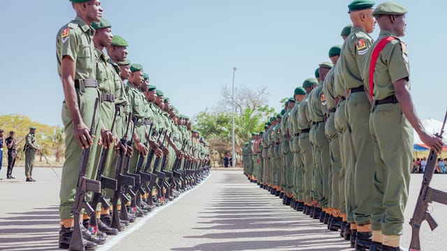 Soldiers in green uniforms stand in formation during a military parade, holding firearms under a clear sky.