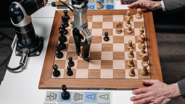 A robotic arm playing chess against a human hand on an electronic chessboard indoors.