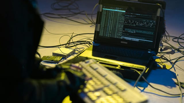 A person typing on a keyboard connected to a laptop showing code, surrounded by wires.