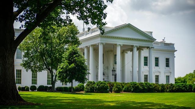 The White House framed by trees and greenery, in Washington, D.C., under a bright sky.