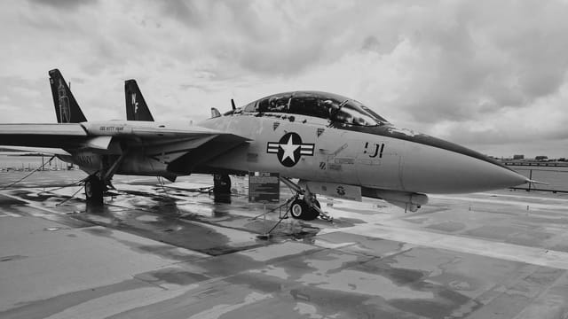 Dramatic grayscale image of a military fighter jet on a wet tarmac under cloudy skies.